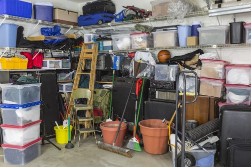 A cluttered storage area filled with various household rubbish and clutter, including multiple plastic storage boxes stacked on metal shelving and on the floor, some with transparent lids showing contents inside. Several gardening tools and cleaning equipment, such as brooms, mops, and buckets in shades of blue, yellow, and terracotta are scattered across the area, with some leaning against the boxes. A wooden ladder is positioned upright among the clutter, partially obscuring a chair placed behind it. The background consists of metal shelves holding miscellaneous items, including plastic containers, bags, and a few larger objects, suggesting an organized but crowded space used for storing household waste and unused items. The scene is well-lit, probably by artificial light, highlighting the various textures of plastic, metal, and wood materials. The overall environment reflects a typical scene for professional rubbish removal services operating in the Shortlands or nearby areas, with a focus on clearing out accumulated household clutter to create a tidy space, as handled by House Clearance Shortlands.